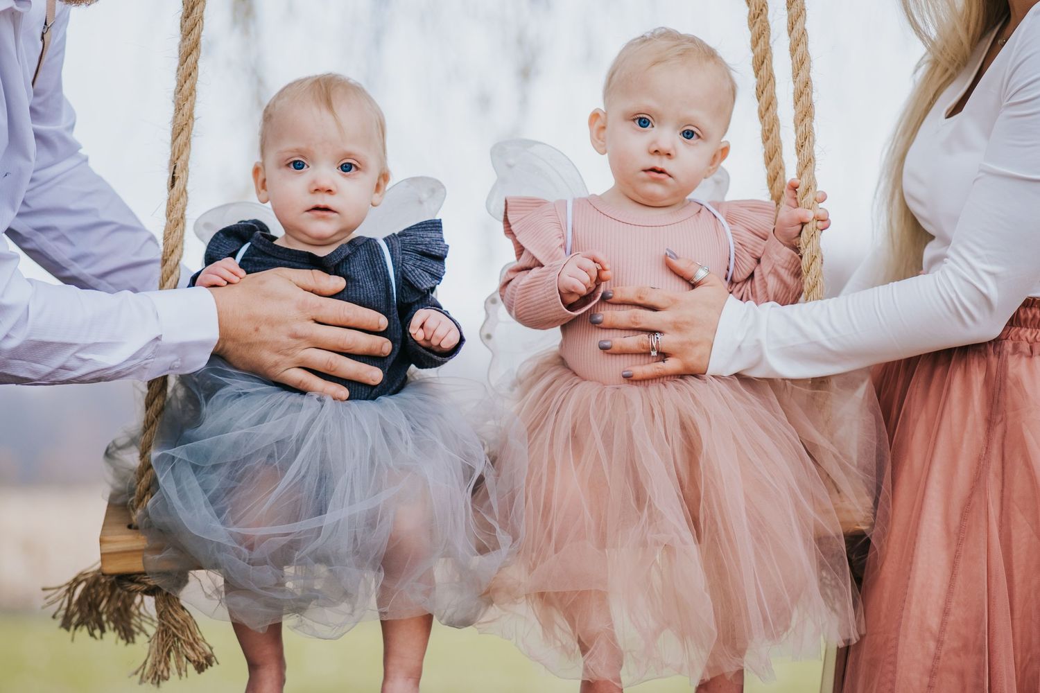 Two baby girls in tutu dresses sitting on a swing, held by adults in a soft outdoor setting.
