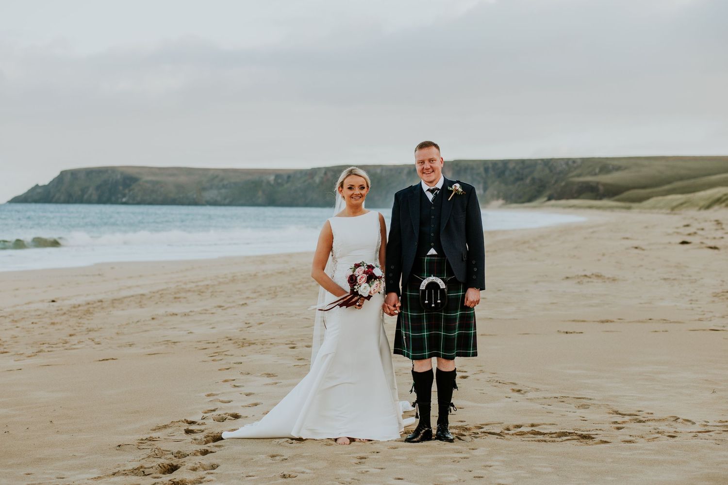 A traditional Scottish wedding couple stands together on a scenic beach with cliffs in the background.