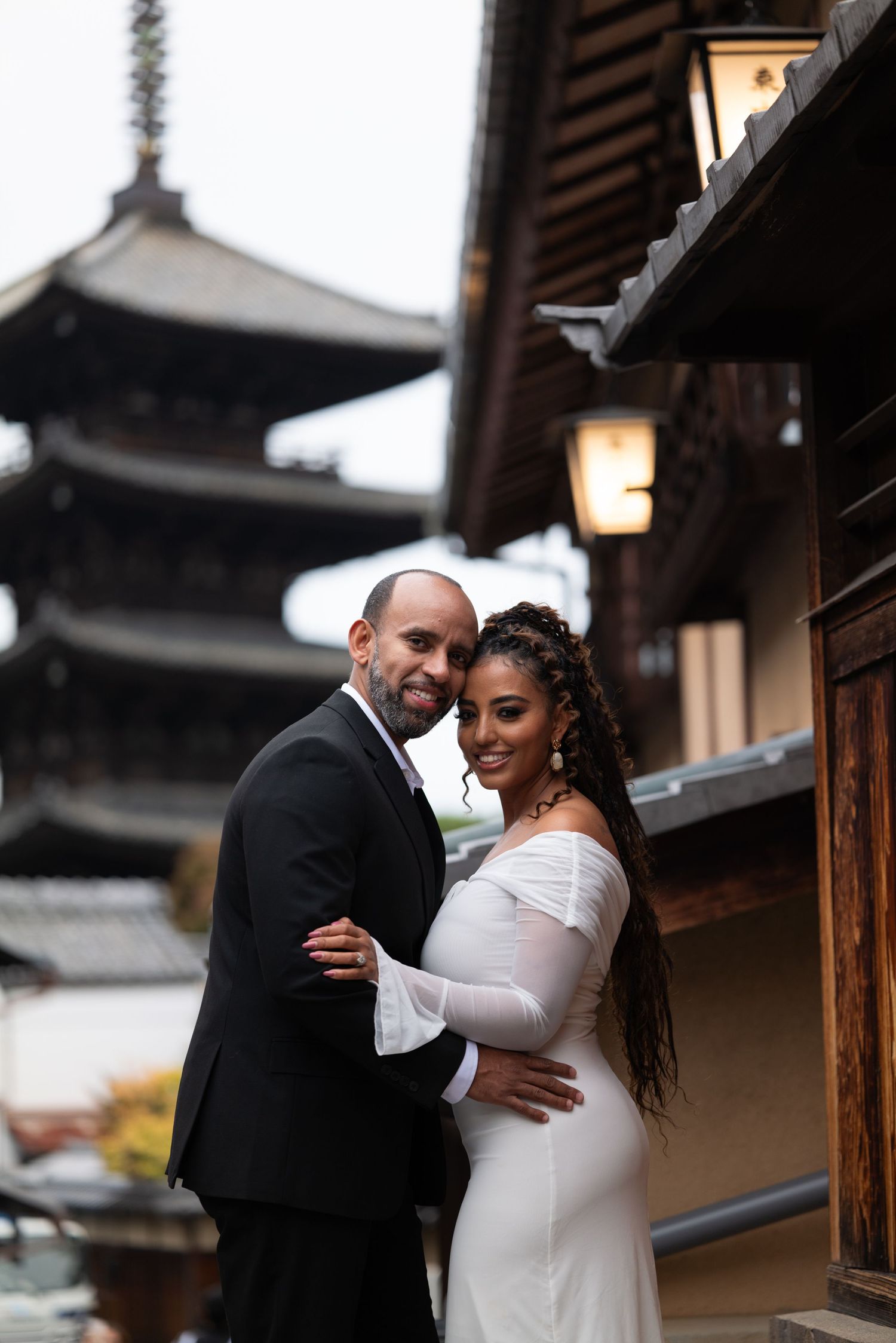 Man in a tan suit kneels on one knee proposing to a woman in a cream dress on a quiet street in front of a historic Kyoto pagoda.