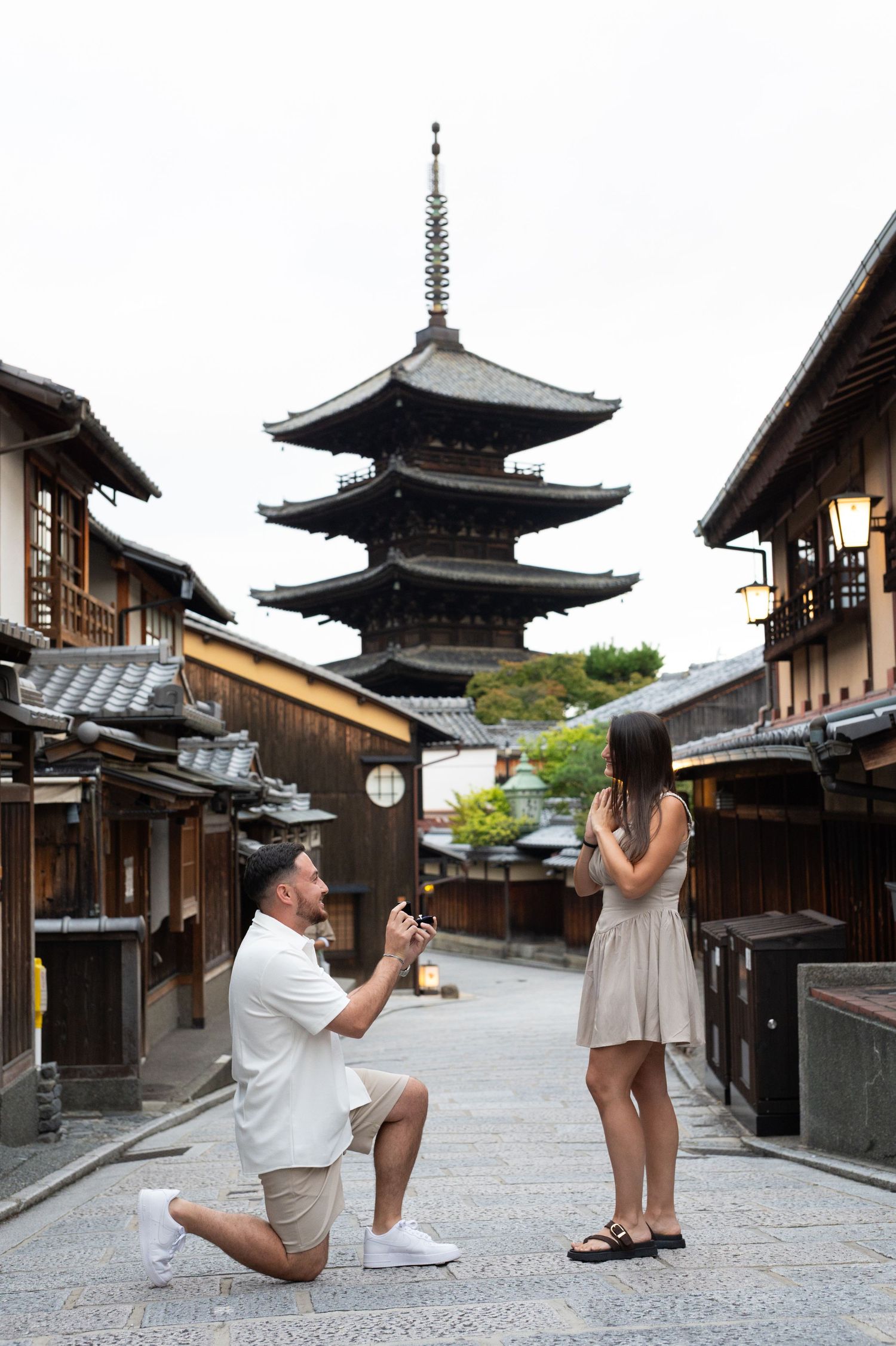 Man kneeling on one knee proposing to a woman on a historic Kyoto street with a traditional wooden pagoda in the background.