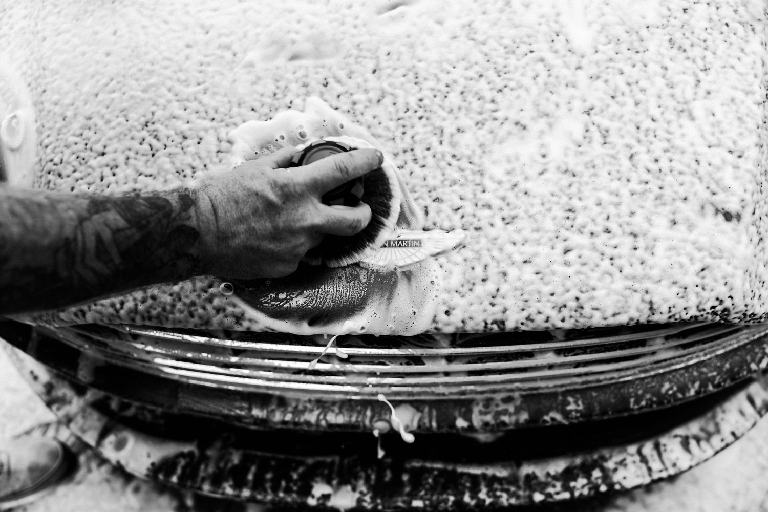 Tattooed hand scrubbing a soapy car hood with a sponge during a car wash in black and white.