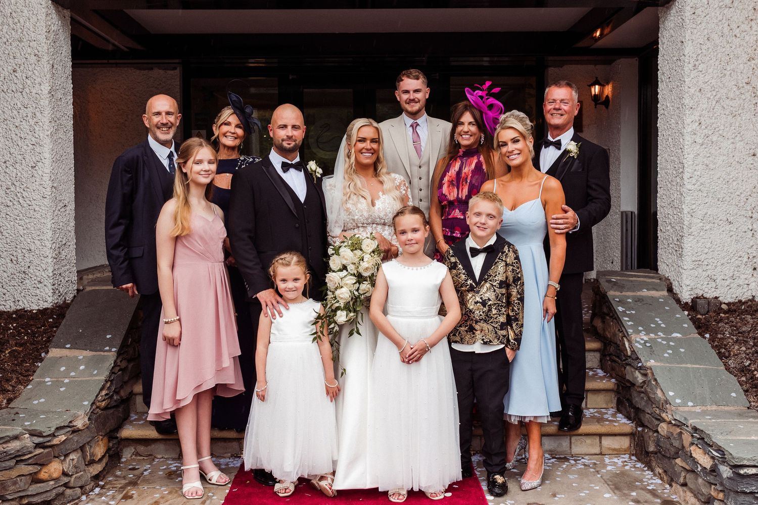 Wedding party poses before rustic wooden doors, bride and groom centered, attendants in gold, orange, and purple gowns with tan suits.