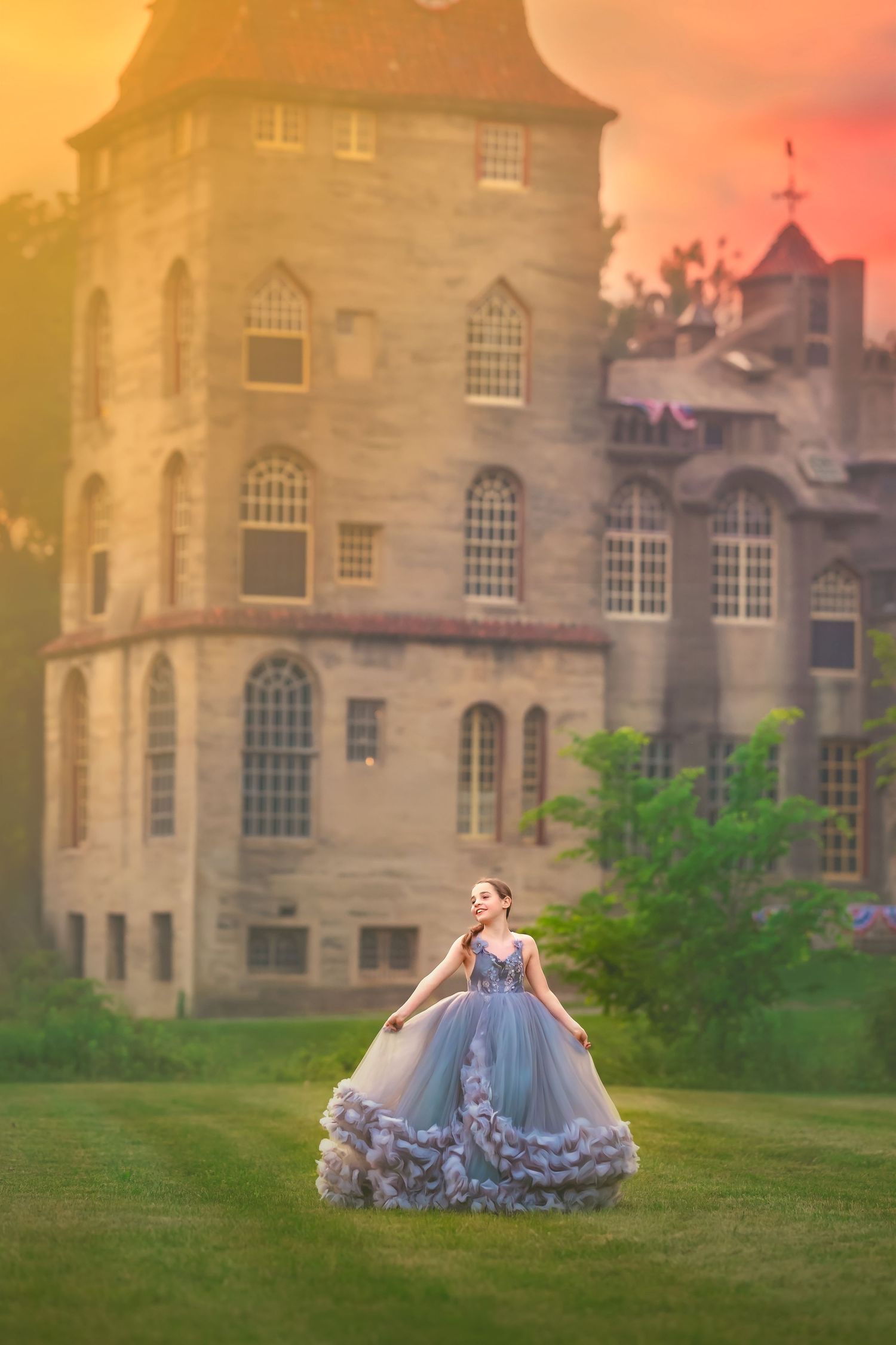 A woman in a flowing blue ball gown stands on green grass before a grand stone castle at golden sunset.