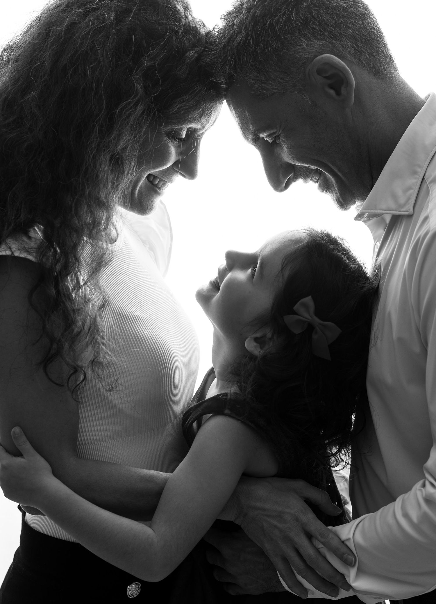 A bearded father and smiling mother tenderly hold their laughing baby together in warm backlit light.