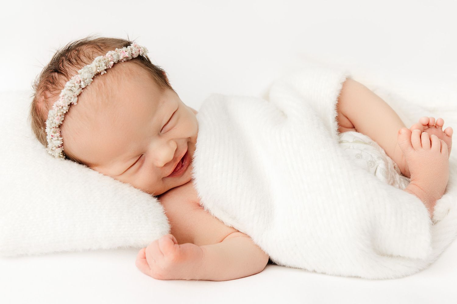Peaceful newborn baby sleeping on white blanket, curled up with hands tucked under chin in serene portrait.