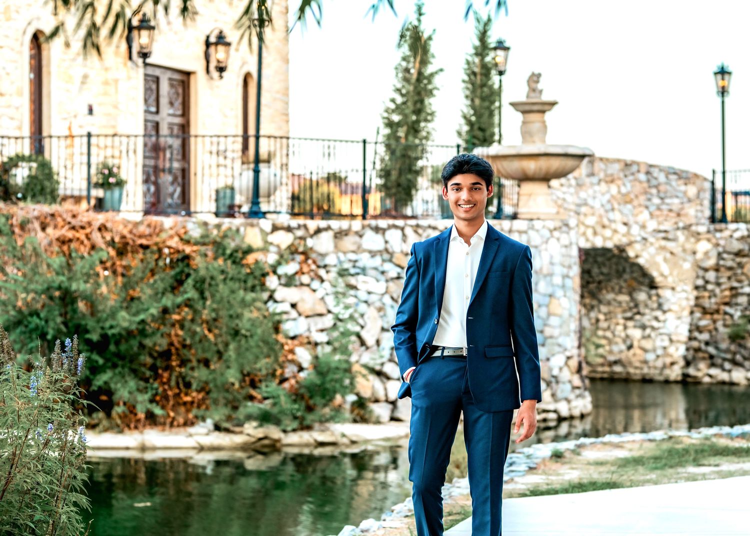 Young man in blue suit sitting on stone steps outdoors, smiling confidently near a scenic water backdrop.