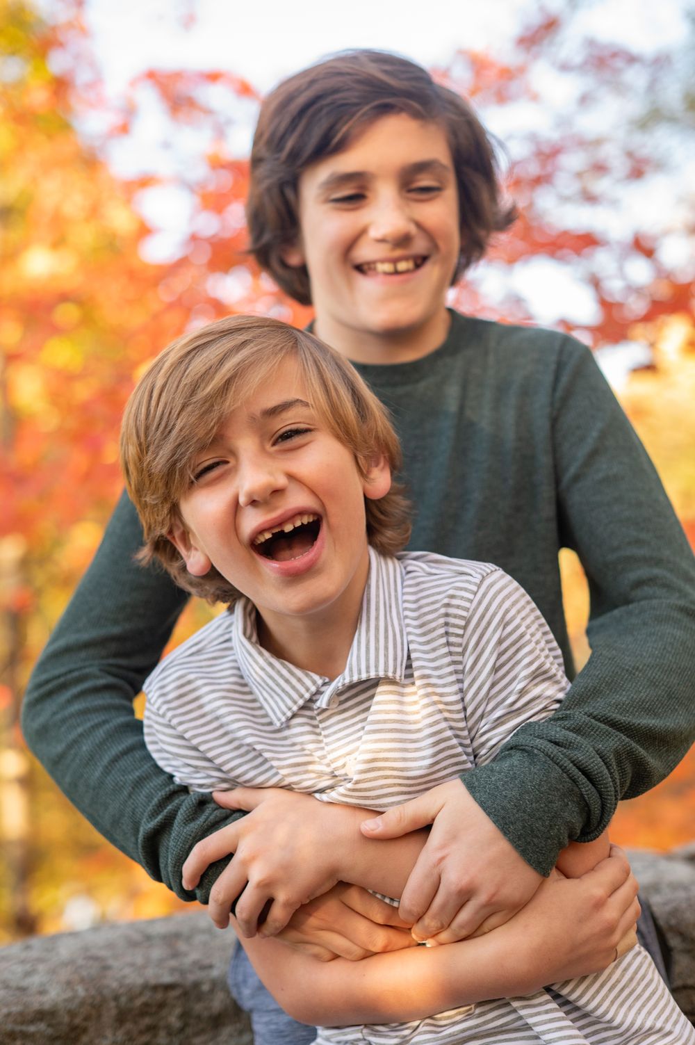 Two laughing boys embrace outdoors among vibrant autumn foliage, capturing a joyful sibling moment in fall.