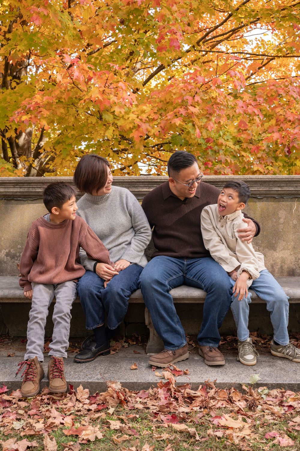 Family of four sitting on a stone bench surrounded by colorful autumn foliage, sharing a joyful moment together.