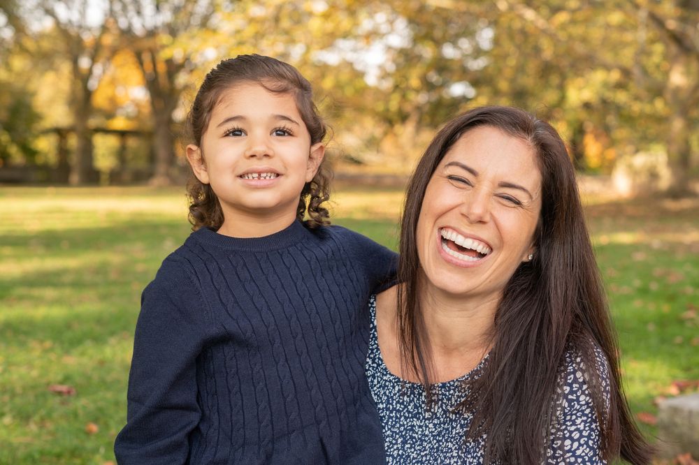 A woman and young girl with braids laugh together outdoors in a park with autumn foliage in the background.
