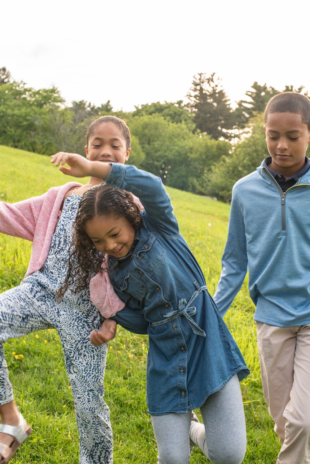Three children playing outdoors on a green grassy hill, laughing and having fun together on a cloudy day.