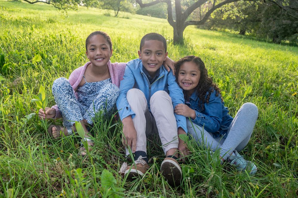 Three smiling children sitting together in a lush green meadow on a sunny day.