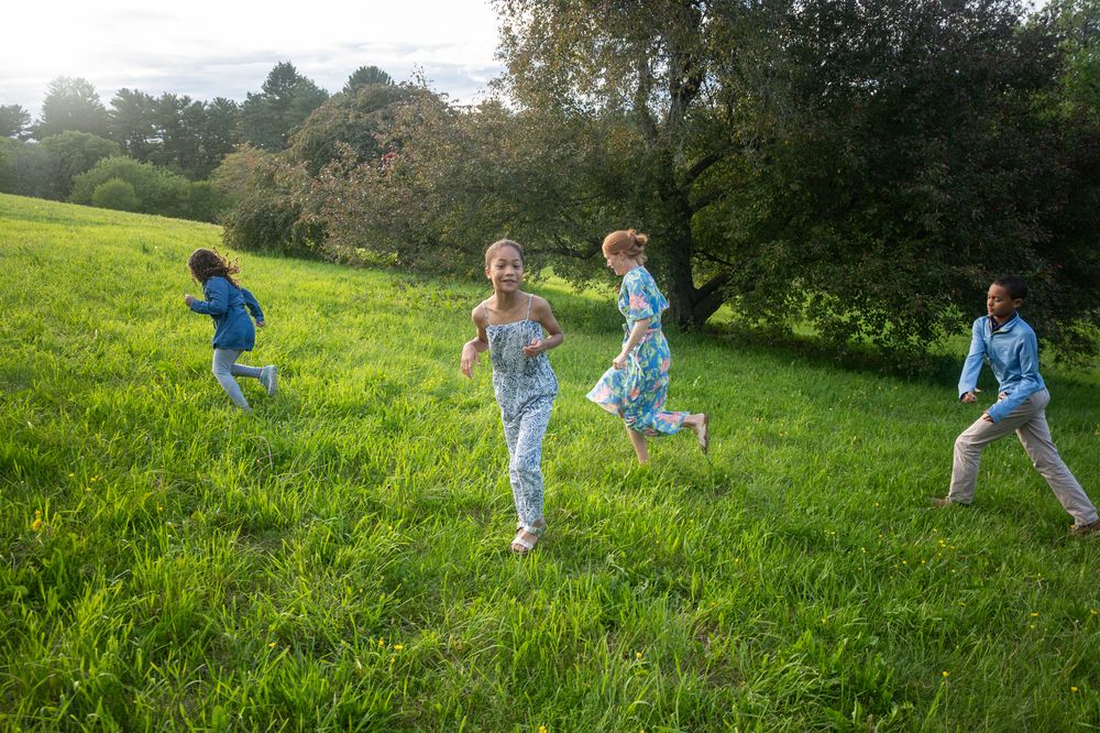 Four children running and playing joyfully on a lush green grassy field surrounded by trees on a sunny day.