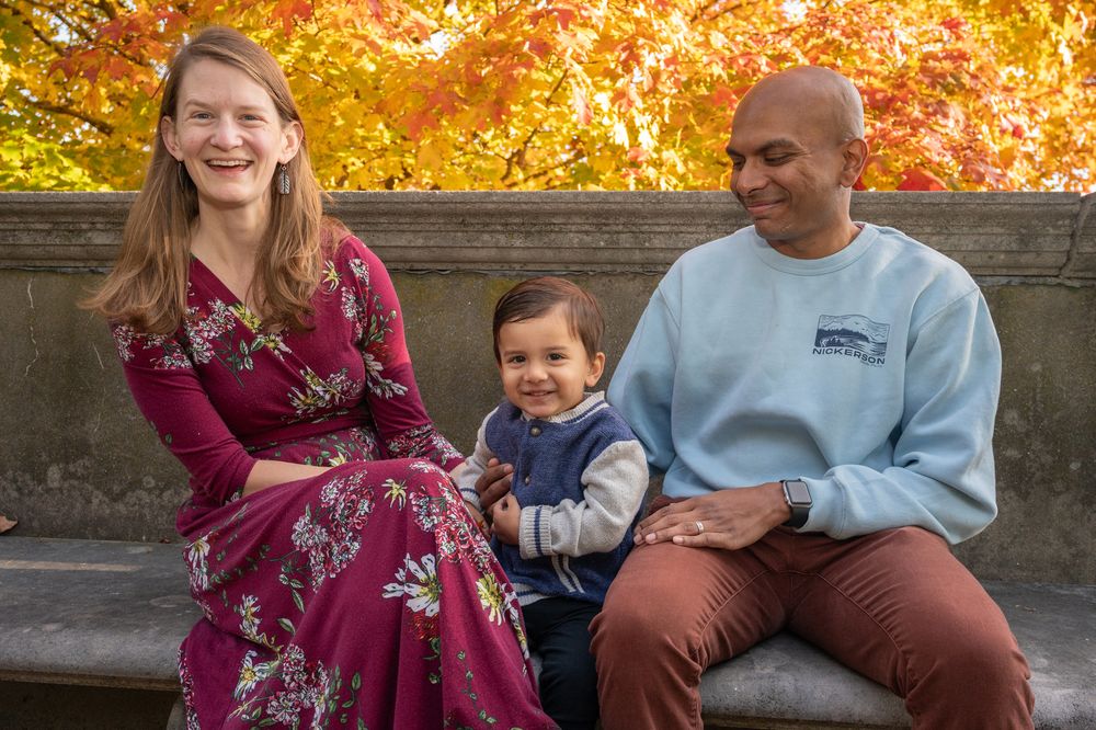 Family of three smiling on a bench surrounded by vibrant autumn foliage in a park setting.