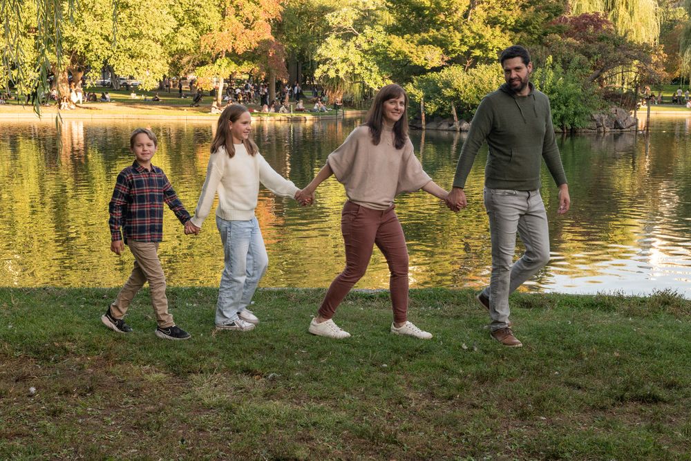 Family of four holding hands and walking along a scenic pond in an autumn park with colorful foliage.