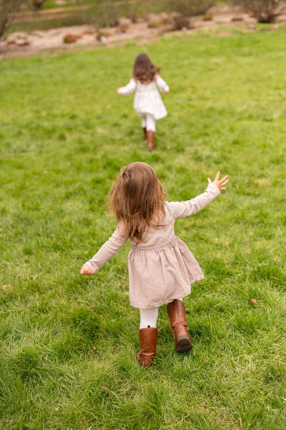 Two young girls in dresses running across a lush green grassy field, viewed from behind.