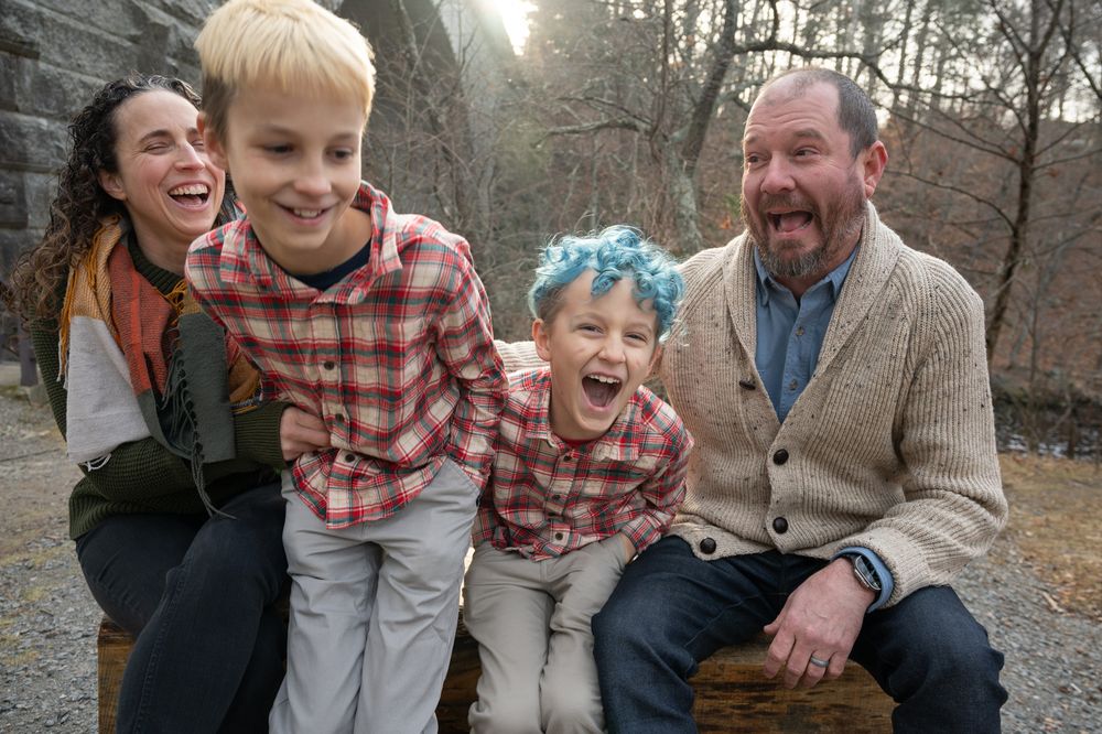 Happy family of four laughing outdoors, with a boy with blue hair sitting on a log in a wooded park setting.