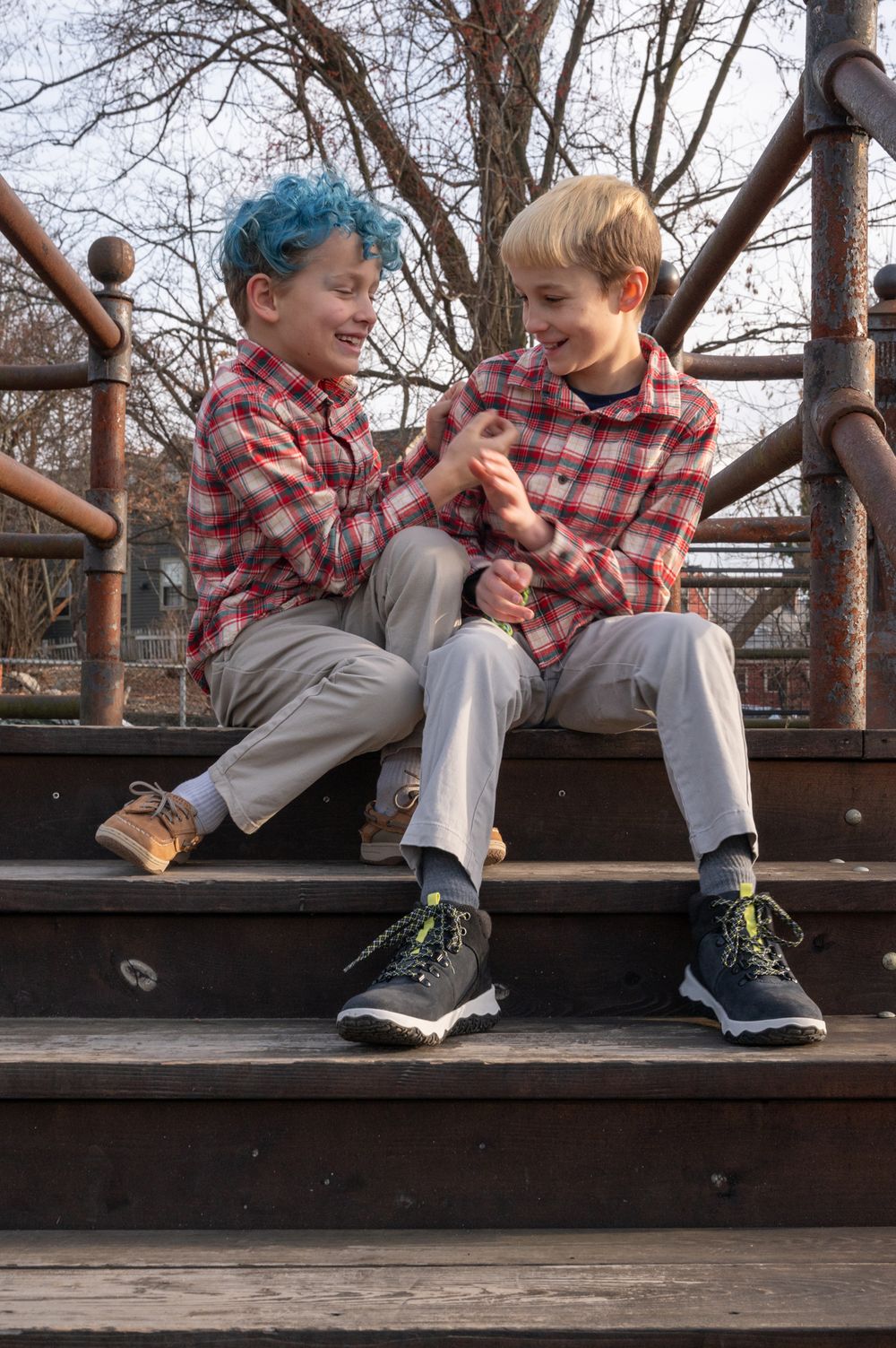 Two boys in matching plaid shirts sitting on wooden steps, smiling and laughing together.