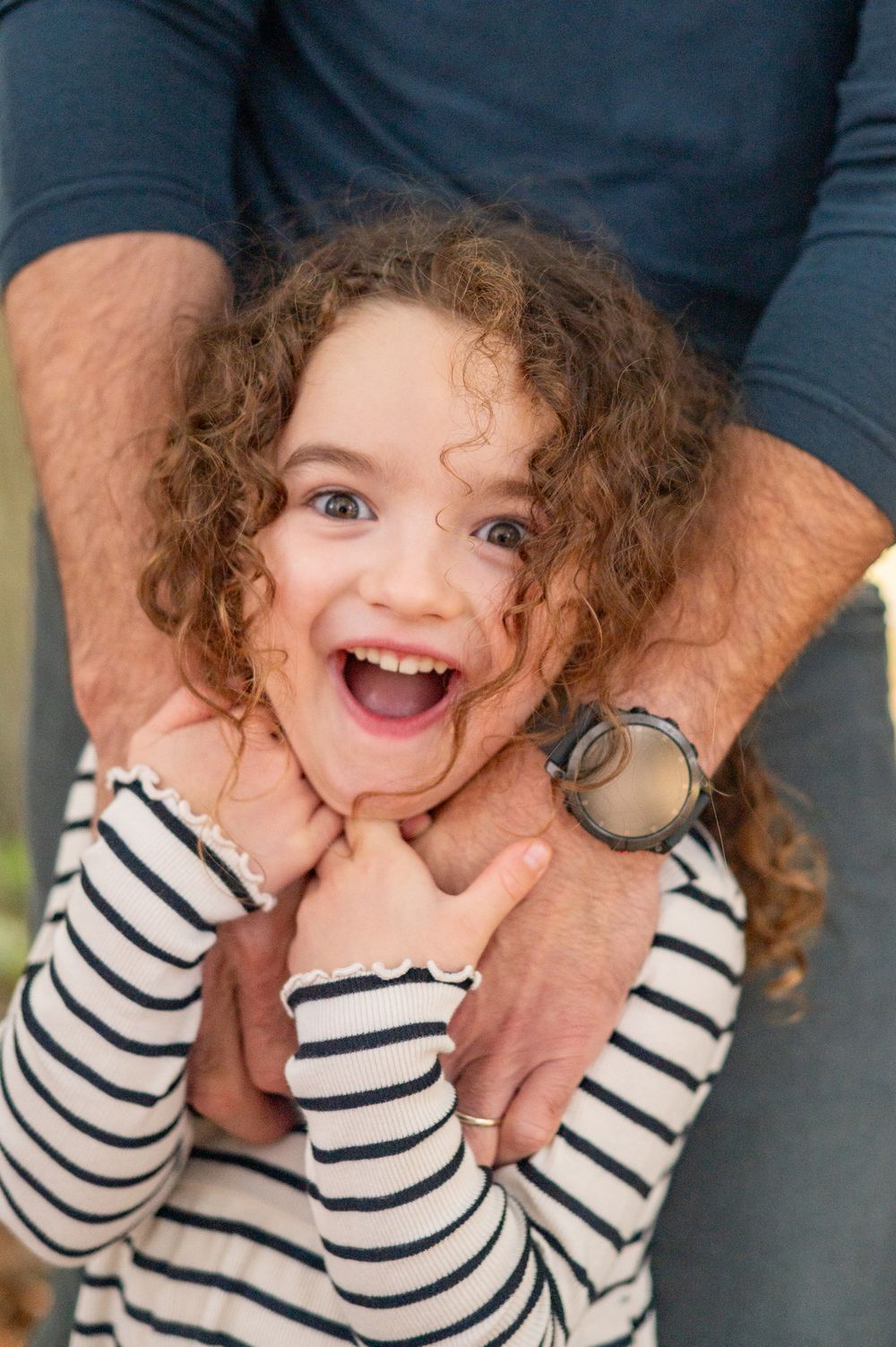 A joyful curly-haired girl laughing while being held by an adult in a fun playful moment outdoors.