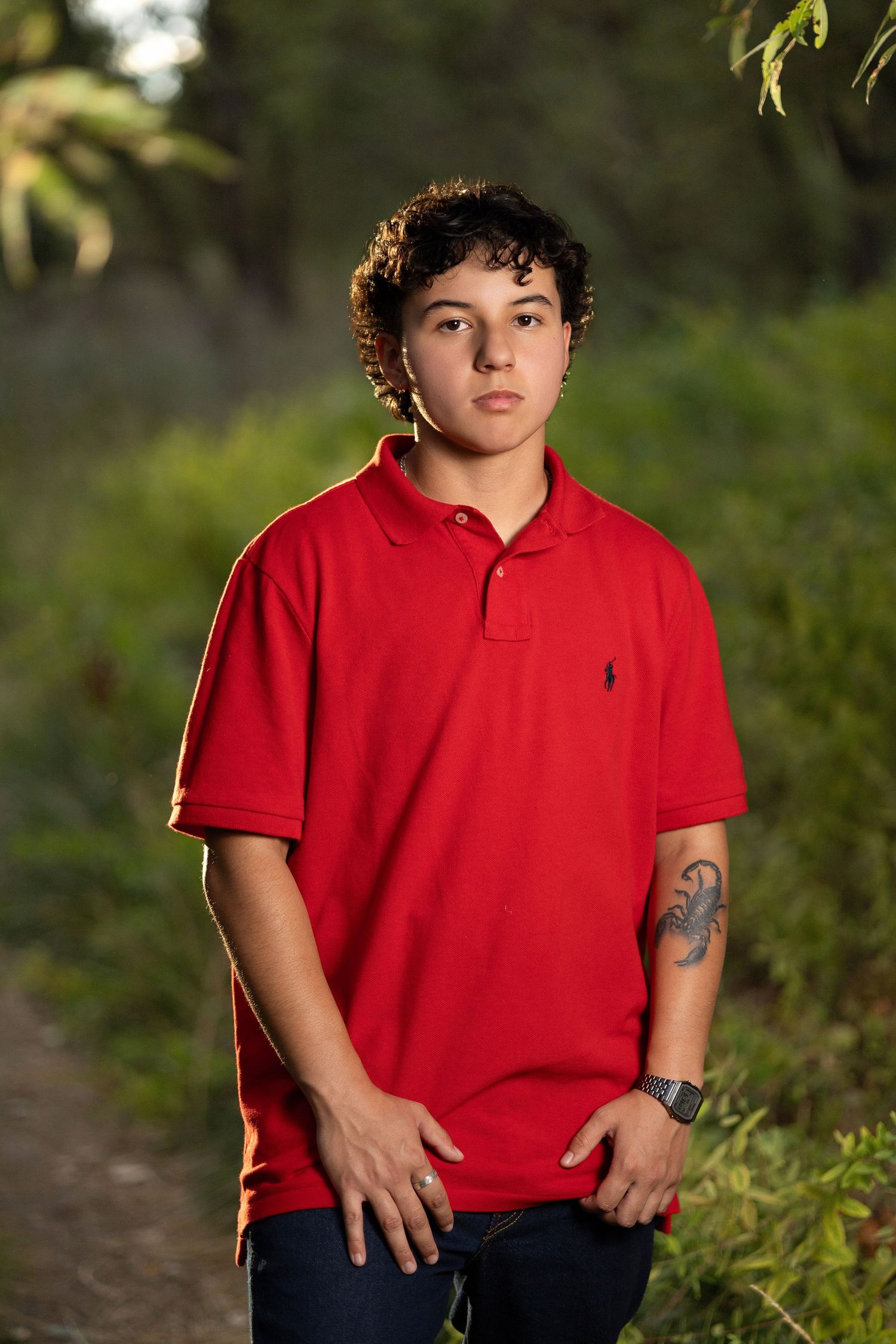 Teen boy in red Ralph Lauren polo shirt with arm tattoo posing outdoors on a nature path.