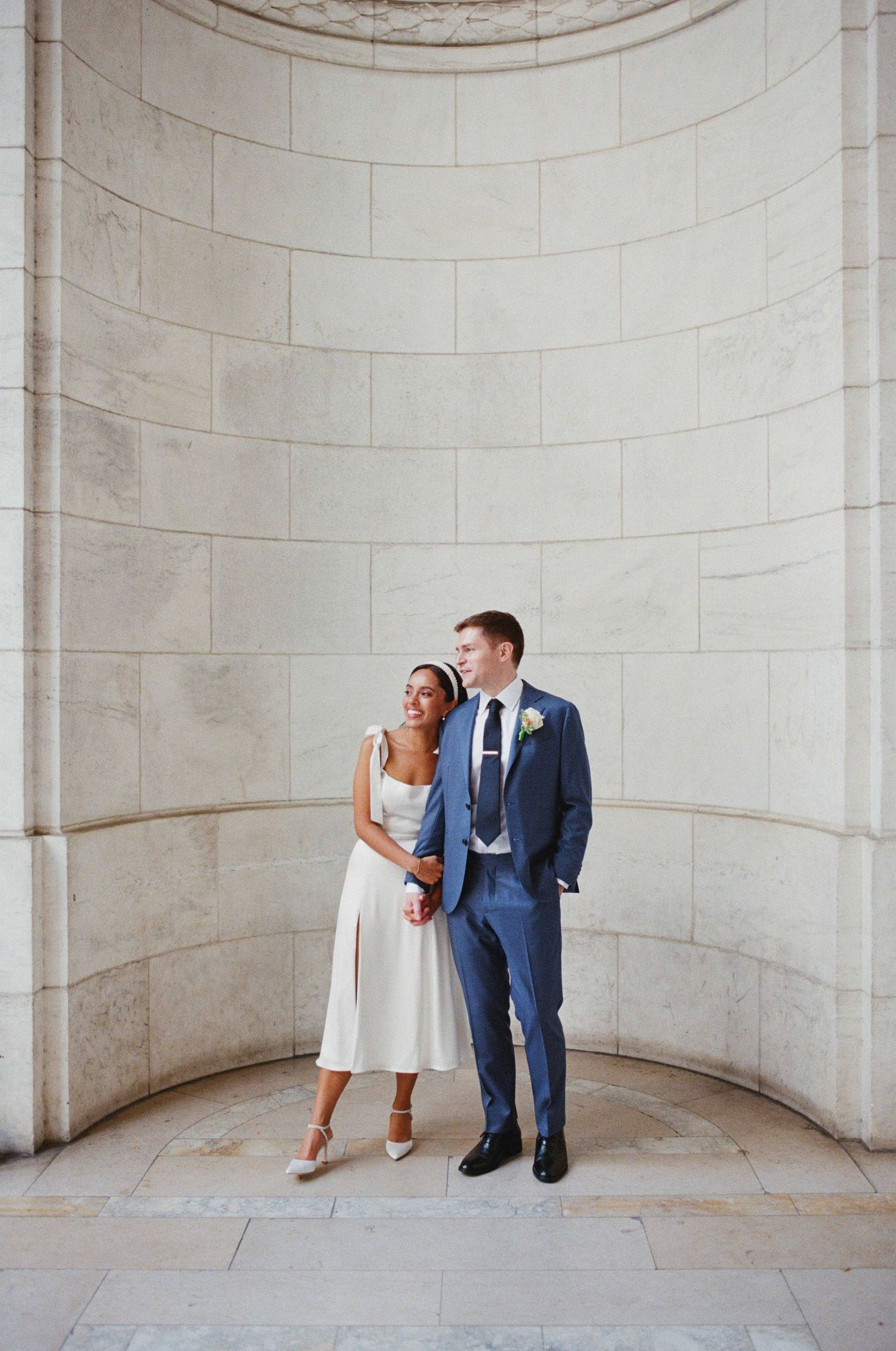 Bride in white midi dress and groom in blue suit pose together in elegant stone archway for wedding portrait.
