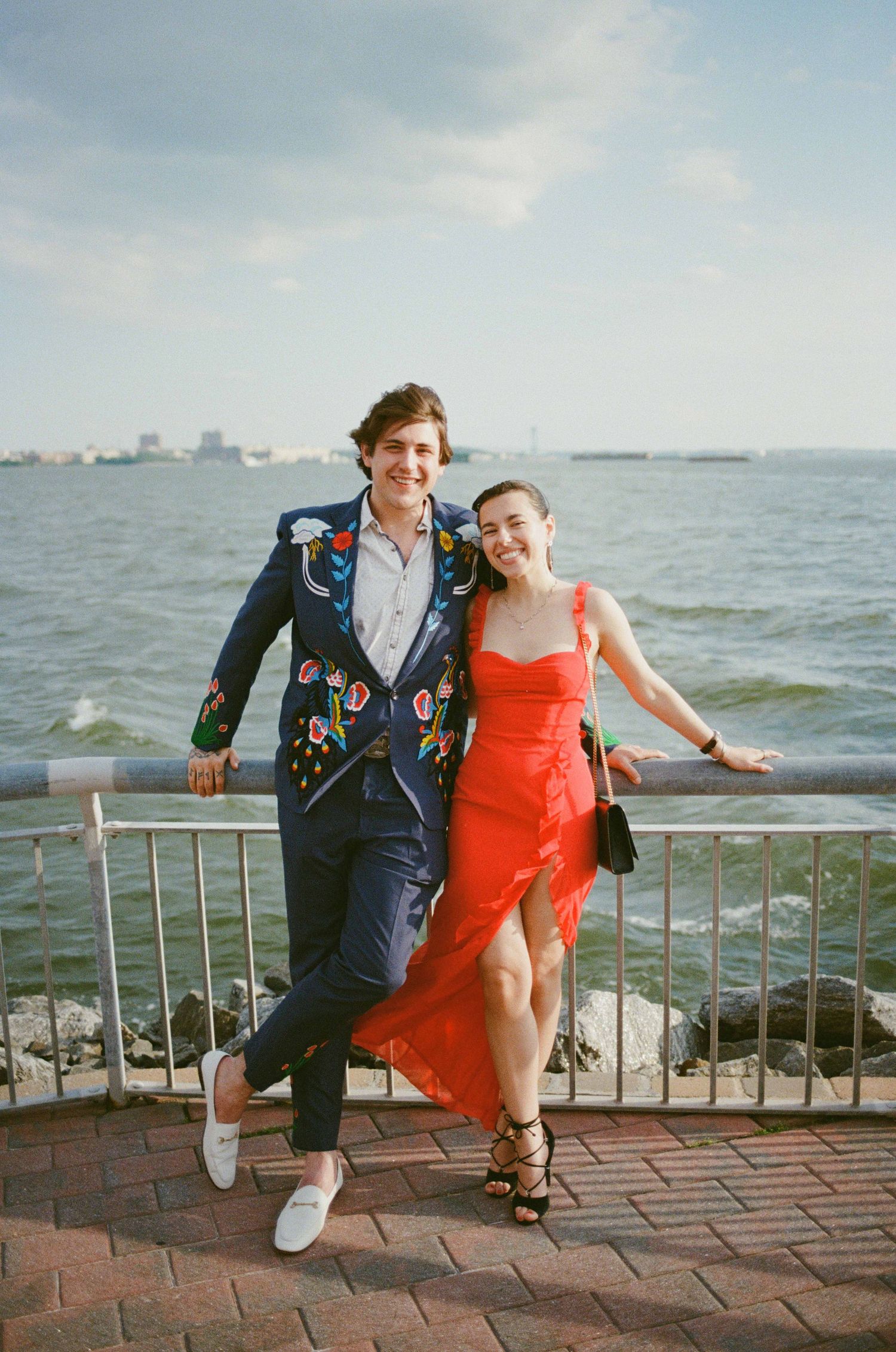 Couple posing by waterfront railing, man in floral blazer and woman in red dress, with ocean backdrop.