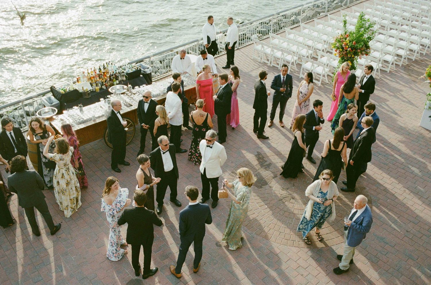 Aerial view of elegantly dressed wedding guests mingling on a waterfront terrace during an outdoor cocktail reception.