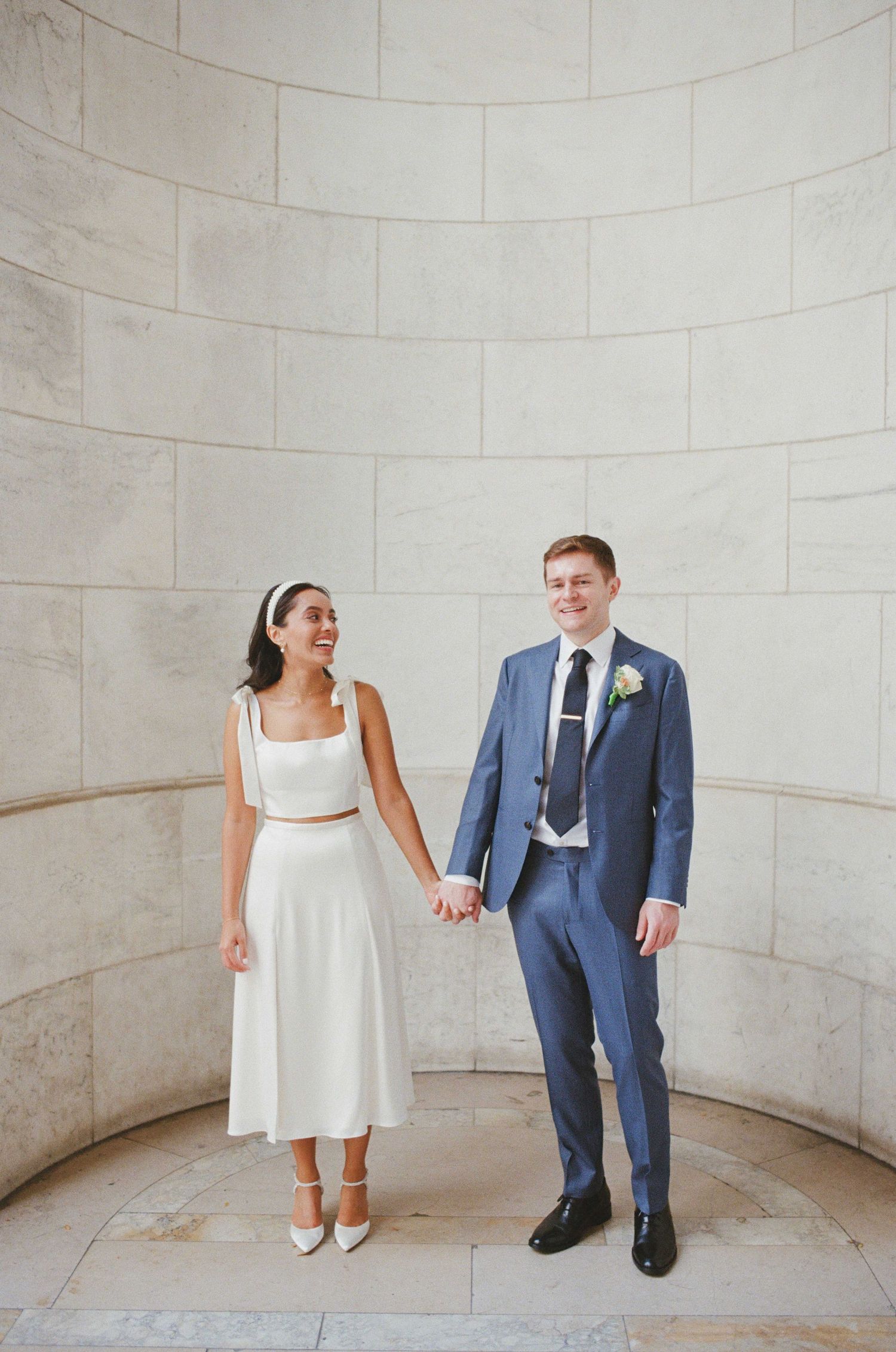 A bride in a white two-piece midi dress and groom in a blue suit hold hands smiling at an elegant stone venue.