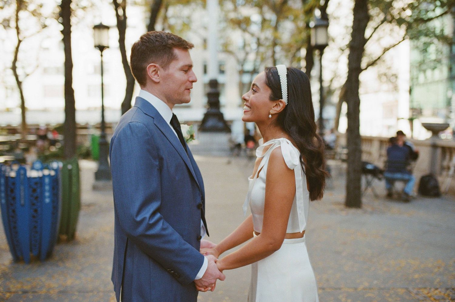 A bride and groom hold hands and gaze at each other in an outdoor plaza, wearing elegant wedding attire.
