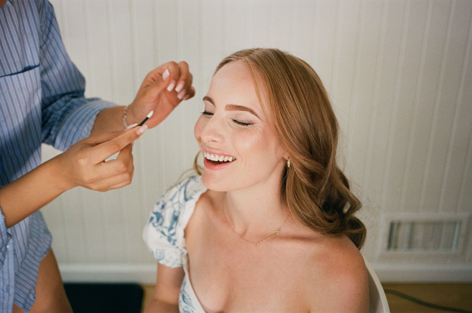 Smiling bride getting makeup applied by a professional artist while sitting in a bright room on her wedding day.