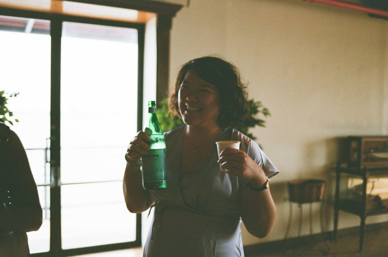 Woman holding a green bottle and cup at an indoor gathering near a bright window.