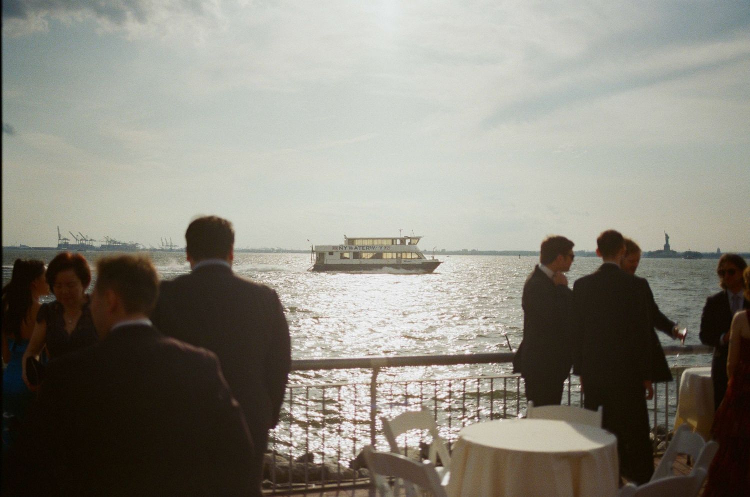 Elegant waterfront event with guests in formal attire, a boat on the water, and a distant statue visible on the horizon.