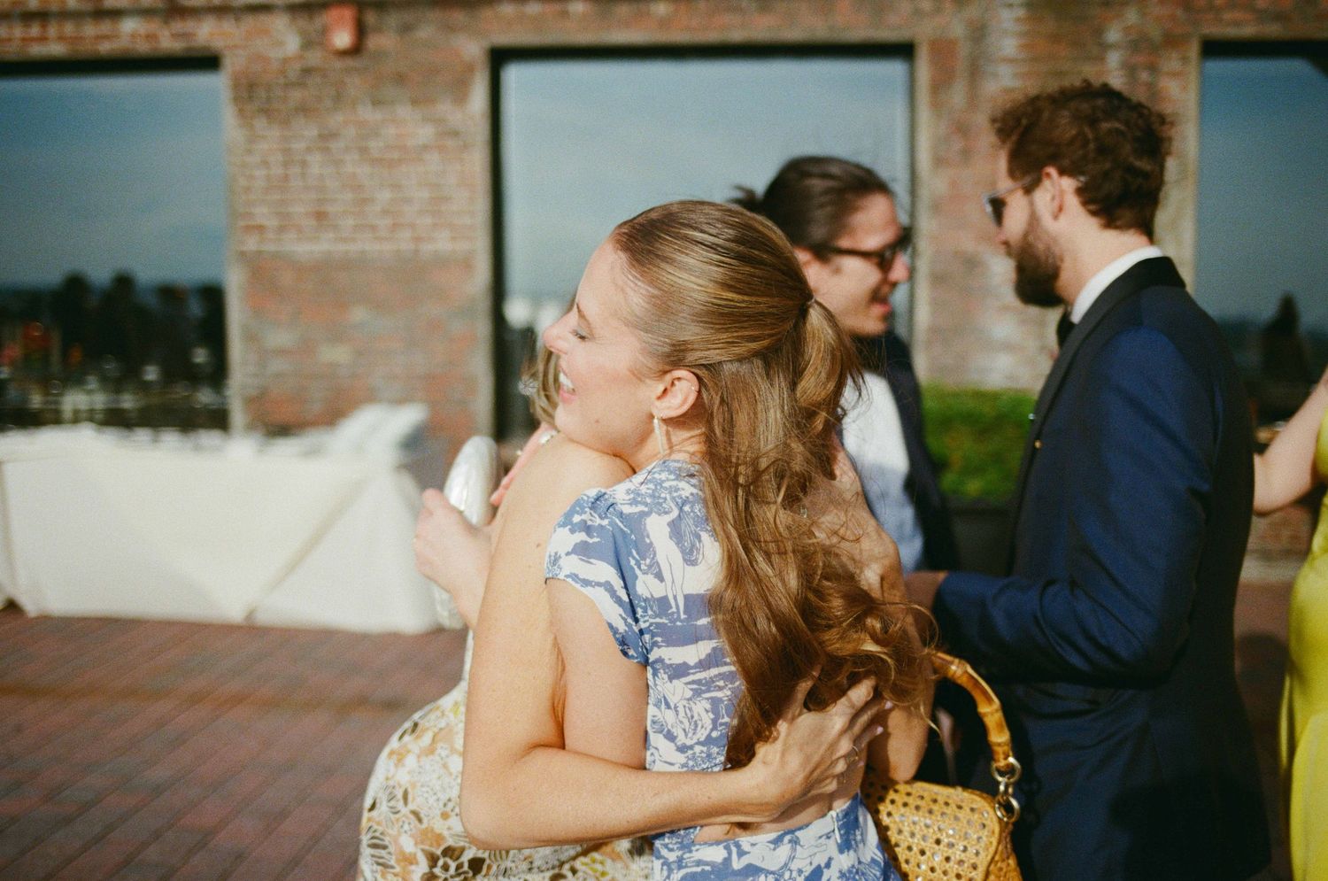 Two women in floral dresses embrace at an outdoor wedding reception near a brick building.