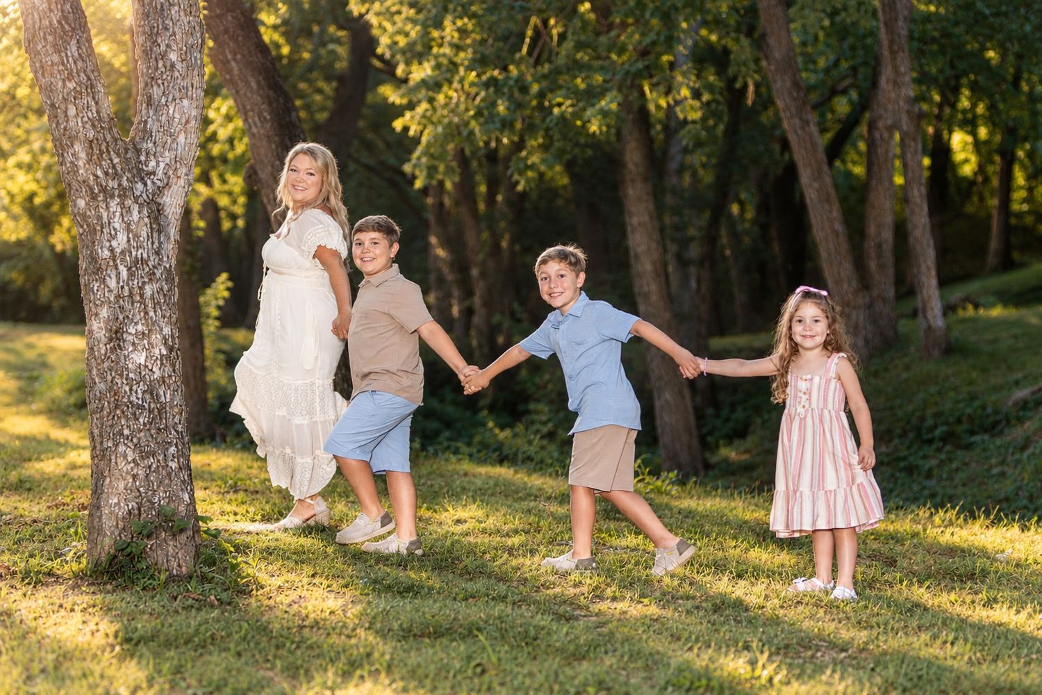 A woman in a white dress holds hands with three children on a sunny green lawn surrounded by trees.