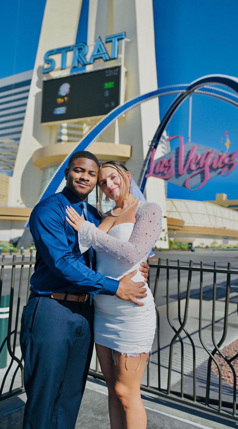 Smiling couple embracing in wedding attire in front of the Strat tower and the Las Vegas sign arch.