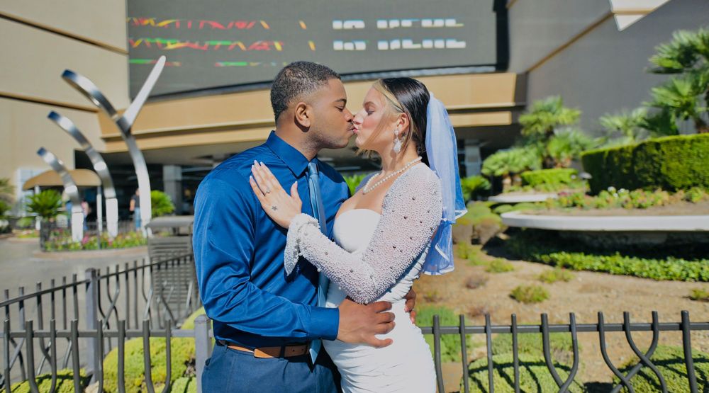 A bride in a lace gown and veil kisses a groom in a blue shirt and tie outdoors near a fenced garden and a modern building.