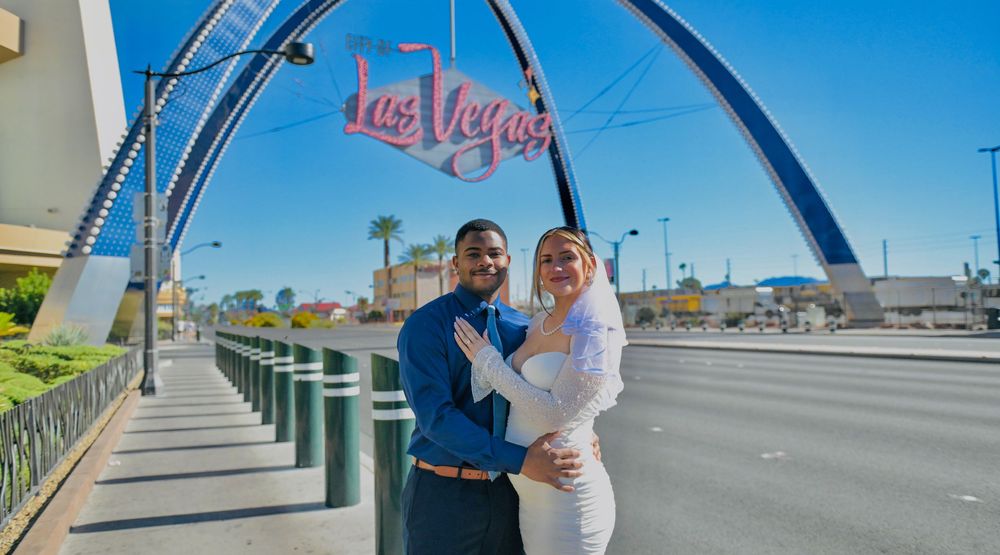 Couple embracing in front of the Welcome to Las Vegas arch sign on a sunny day, the bride in white and the groom in a navy shirt.