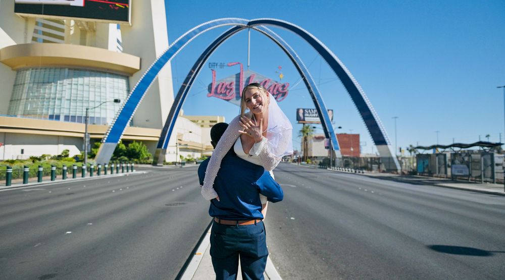Groom carrying a bride in a white veil down an empty Las Vegas street beneath the iconic arches on a sunny day.