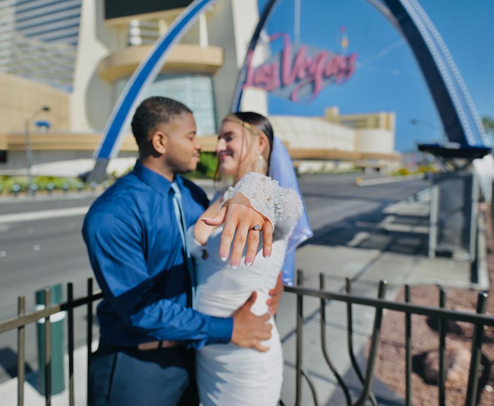 Couple embracing near the Welcome to Las Vegas sign as the woman shows off her new engagement ring on a sunny day.