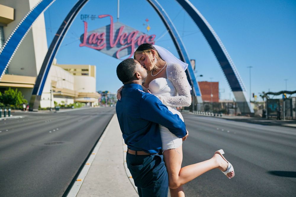Groom in a blue shirt lifts his veiled bride in a white dress as they kiss on a quiet street near the Las Vegas sign and arch.