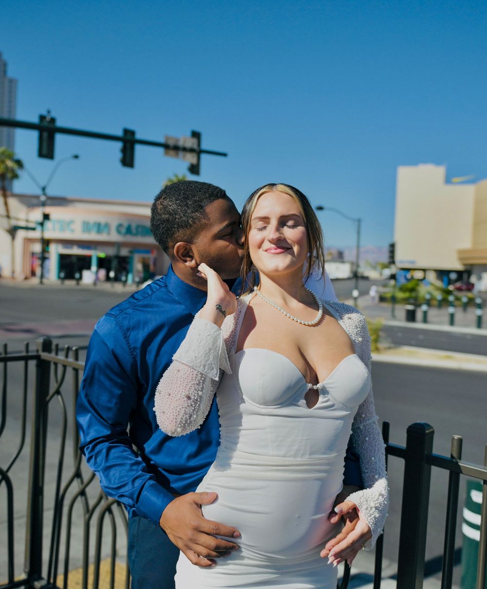 Bride in a white dress and lace sleeves smiles with closed eyes as her groom in a blue shirt kisses her cheek on a Las Vegas street.