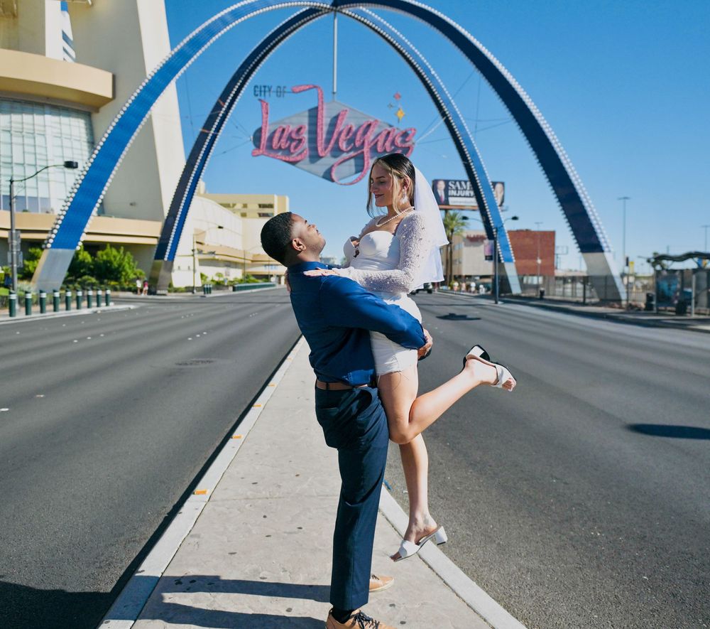 Groom lifting a bride in a white dress and veil near the iconic Las Vegas arches on a sunny Las Vegas Boulevard.