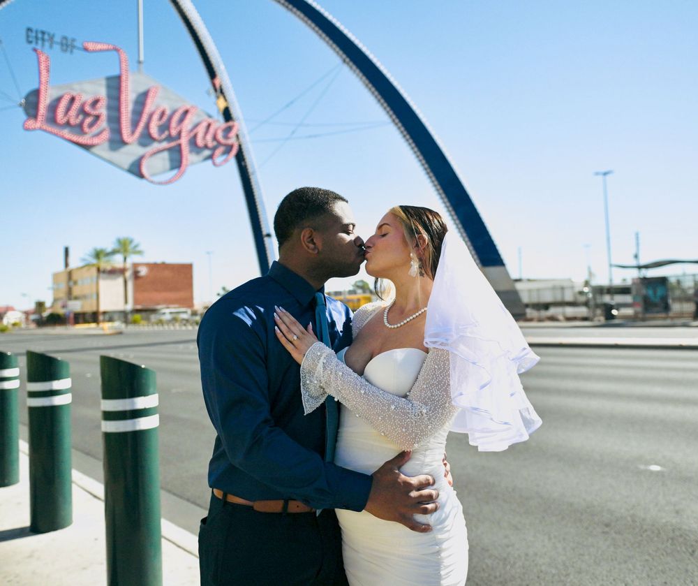 Newlywed couple kisses beneath the Welcome to Las Vegas arch, the bride in a white dress and veil, the groom in a blue shirt.