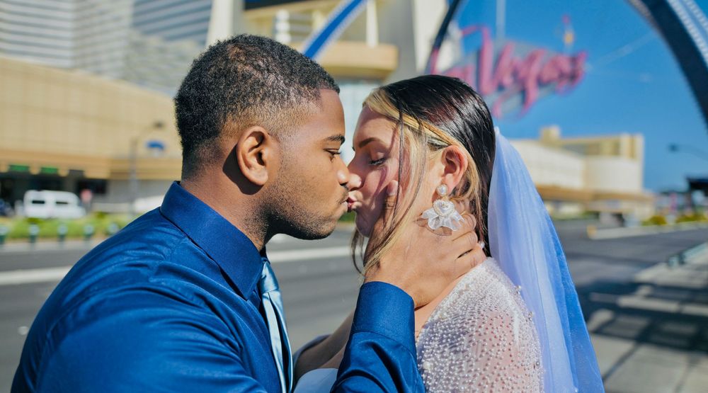 Newlywed couple in wedding attire kissing outdoors on a sunny Las Vegas street, the bride wearing a veil and the groom a blue suit.