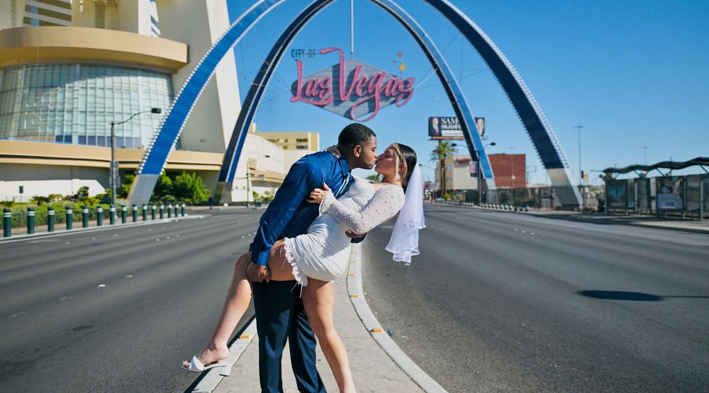 Groom dips his veiled bride in a romantic kiss on a Las Vegas street beneath the iconic arch and sign on a sunny day.