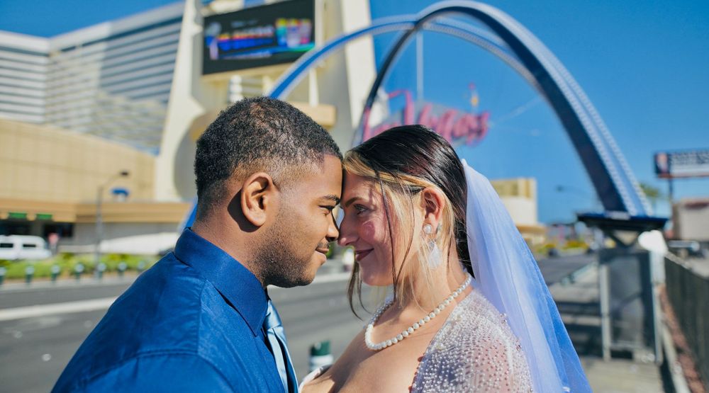 Bride in a veil and pearl necklace touches foreheads with groom in a blue shirt beneath a city arch on a sunny day.