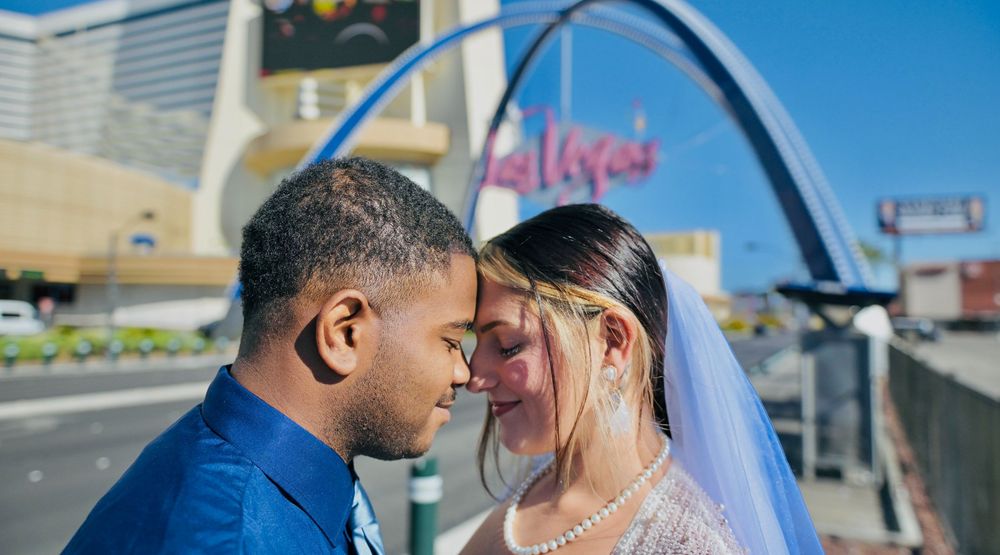 A bride in a veil and pearl necklace touches foreheads with a groom in a blue shirt beneath a blue arch in a sunny city setting.