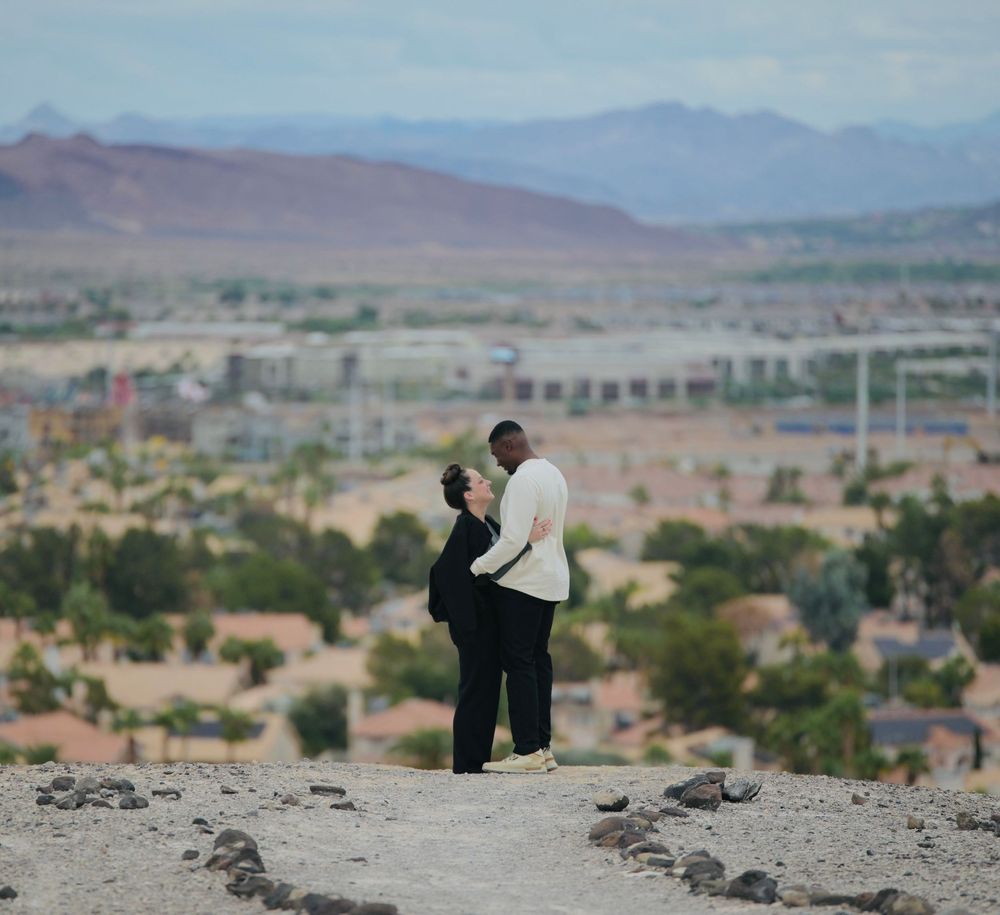 A couple in an embrace on a rocky desert overlook with a sprawling city and distant mountains stretching behind them under a cloudy sky.
