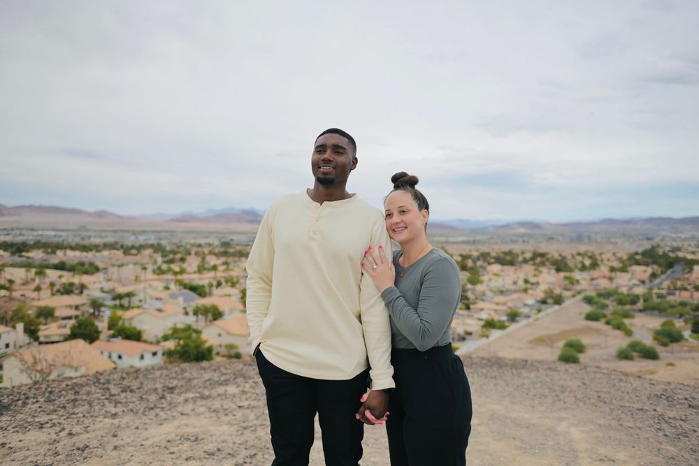 A smiling couple holds hands on a desert hilltop overlooking a sprawling suburban neighborhood with mountains under a cloudy sky.