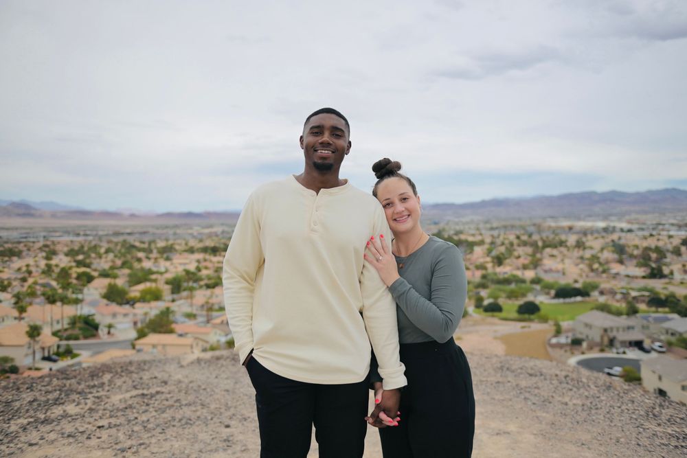 Smiling couple embracing on a rocky hilltop overlooking a sprawling desert city with mountains under a cloudy sky.