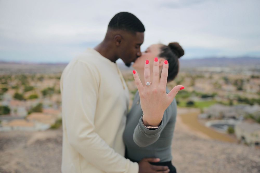 Newly engaged couple kissing as the woman shows off her diamond engagement ring with red nails against a scenic overlook of a desert city.